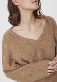 Miękki, teksturowany, brązowy sweter z szerokim dekoltem w kształcie litery V i długimi, luźnymi rękawami. Materiał wydaje się puszysty i ciepły, idealny na chłodniejsze dni.