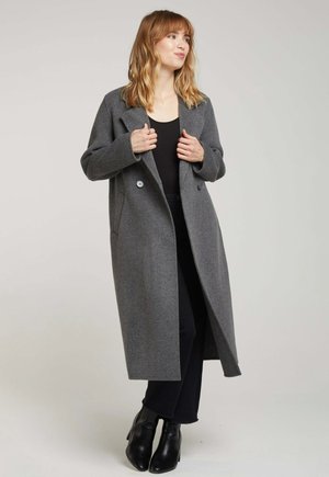 Manteau gris à double boutonnage avec une silhouette longue, en laine, présentant des revers crantés et deux poches avant. Associé à un pantalon noir et des bottes.