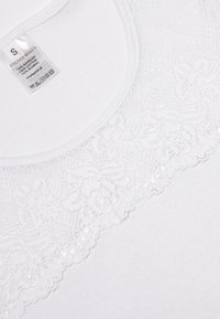Biała bawełniana koszulka z okrągłym dekoltem, ozdobiona delikatnym koronkowym wykończeniem o kwiatowych wzorach oraz subtelnym po łyskiem. Widoczna metka wewnątrz.