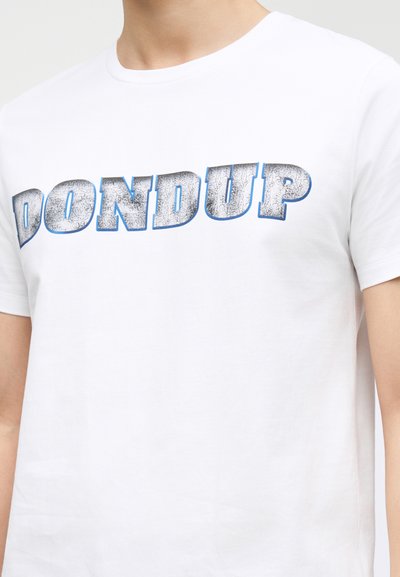 Άτομο που φοράει λευκό μπλουζάκι με μεγάλα, ραγισμένα ασημένια γράμματα περιγραμμένα με μπλε που σχηματίζουν τη λέξη "DONDUP" στο στήθος.