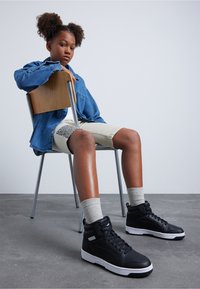 Zwarte high-top sneakers met witte accenten, een denim overhemd met knopen, shorts met een gemusterde zijkant en grijze sokken, zittend op een houten stoel.