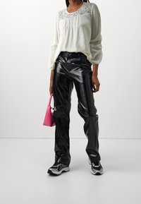 Blusa bianca a maniche lunghe con dettagli in pizzo, abbinata a pantaloni neri lucidi. Tiene una piccola borsa rosa e indossa sneakers bianche e nere.