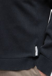 Gros plan d'un pull texturé bleu marine avec une petite étiquette blanche sur le côté et une main partiellement dans la poche d'un pantalon beige.