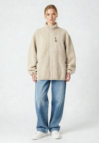 Giacca in pile beige con colletto alto, maniche lunghe, cerniera frontale e tasche laterali. Abbinata a jeans a gamba dritta azzurri chiaro e sneakers bianche.