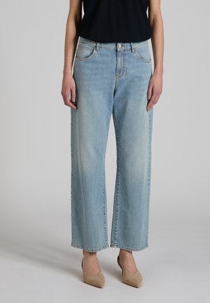 Kobieta w jasnoniebieskich dżinsach z szerokimi nogawkami, czarnym topie bez rękawów i beżowych szpilkach z czubkiem, stoi na jednolitym tle.