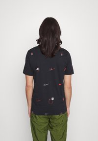 Camiseta de algodón negra con varios logotipos de Nike en rojo y blanco. Mangas cortas, corte regular, con una textura suave. Combinada con pantalones verdes.