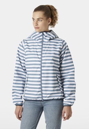Vrouw draagt een blauw-wit horizontaal gestreepte capuchonjas met rits en lichtblauwe spijkerbroek, kijkend naar links.