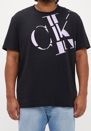 Hombre con camiseta negra con un diseño de grandes letras fragmentadas de color lila claro en la parte superior frontal, combinado con vaqueros azul claro.