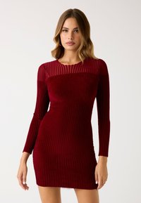 Abito rosso a coste con maniche lunghe e scollatura trasparente. Il tessuto è morbido e testurizzato. Silhouette aderente con lunghezza mini.