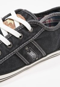 Tmavě šedé plátěné tenisky s bílými tkaničkami, bílým prošíváním, kostkovanou vnitřní podšívkou a malou kovovou značkou na boku blízko podrážky.