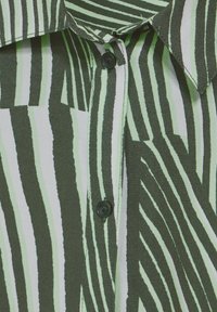 Zielono-biała bluzka w paski z kołnierzykiem, z zapięciem na guziki i teksturowanym materiałem. Paski różnią się szerokością i wzorem.
