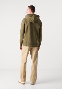 Olijfgroene hoodie met een grote capuchon, geribbelde boorden en zoom. Gecombineerd met lichtbeige broek en witte schoenen met bruine zolen.