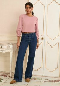 Helles rosa Blusenshirt mit Puffärmeln und Raffung, kombiniert mit dunkelblauen Schlagjeans und braunen Schuhen. Weißer dekorativer Hintergrund.