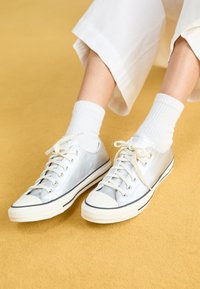 Zapatillas de lona plateadas con suelas y cordones de goma blanca, con un acabado brillante y una franja negra en contraste a lo largo del borde.