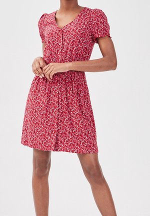 Femme portant une robe rouge à fleurs à manches courtes avec des boutons sur le devant, debout devant un fond clair uni.