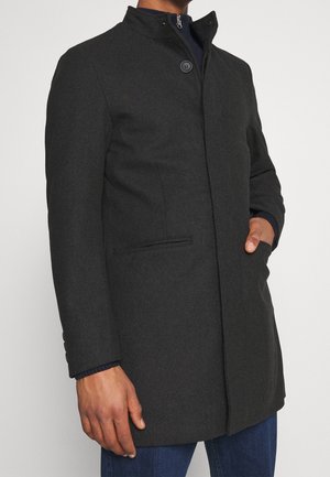 Homme portant un manteau en laine gris foncé boutonné, une main dans la poche et un jean bleu sur un fond uni.