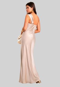 Robe sans manches en satin avec une épaule drapée et un dos nu. Couleur beige clair, design long jusqu'au sol et éclat subtil. Accent en métal doré.