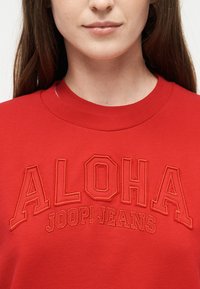 Κοντινή λήψη ενός ατόμου που φοράει κόκκινο φούτερ με το κέντημα "ALOHA JOOP! JEANS" στο μπροστινό μέρος.