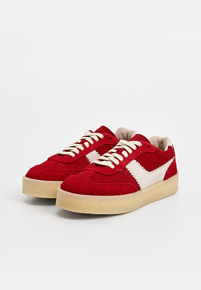 Baskets en daim rouge avec des accents beige, présentant un logo blanc et un design classique à lacets. Semelle épaisse et texturée pour le confort et le soutien.