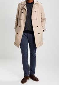 Manteau beige avec des boutons marron, porté sur un pull à motifs navy. Associé à un pantalon navy et des bottines marron.