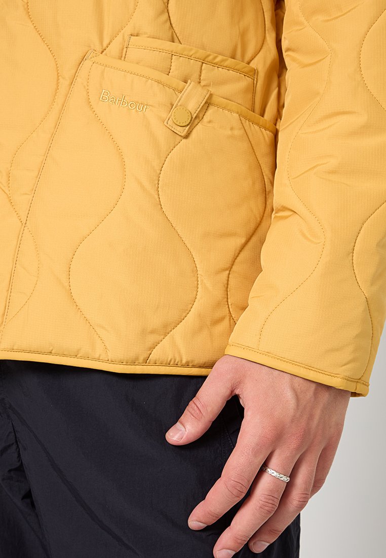 Gelbe gesteppte Jacke mit einem strukturierten Wellenmuster, seitlicher Tasche mit Knopfverschluss und einer glatten Stoffoberfläche.