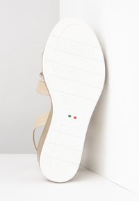 Sandale beige avec une semelle en caoutchouc blanc, présentant une surface texturée. Le dessous montre un logo et un design lisse avec des motifs d'adhérence.