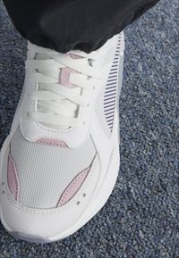 Zapatilla blanca con un upper texturizado, acentos en rosa y rayas moradas. Suela gruesa, punta redondeada y diseño tradicional de cordones.