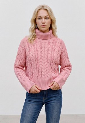 Jeune femme aux cheveux blonds portant un pull à col roulé en maille torsadée rose et un jean bleu, debout devant un fond clair uni.