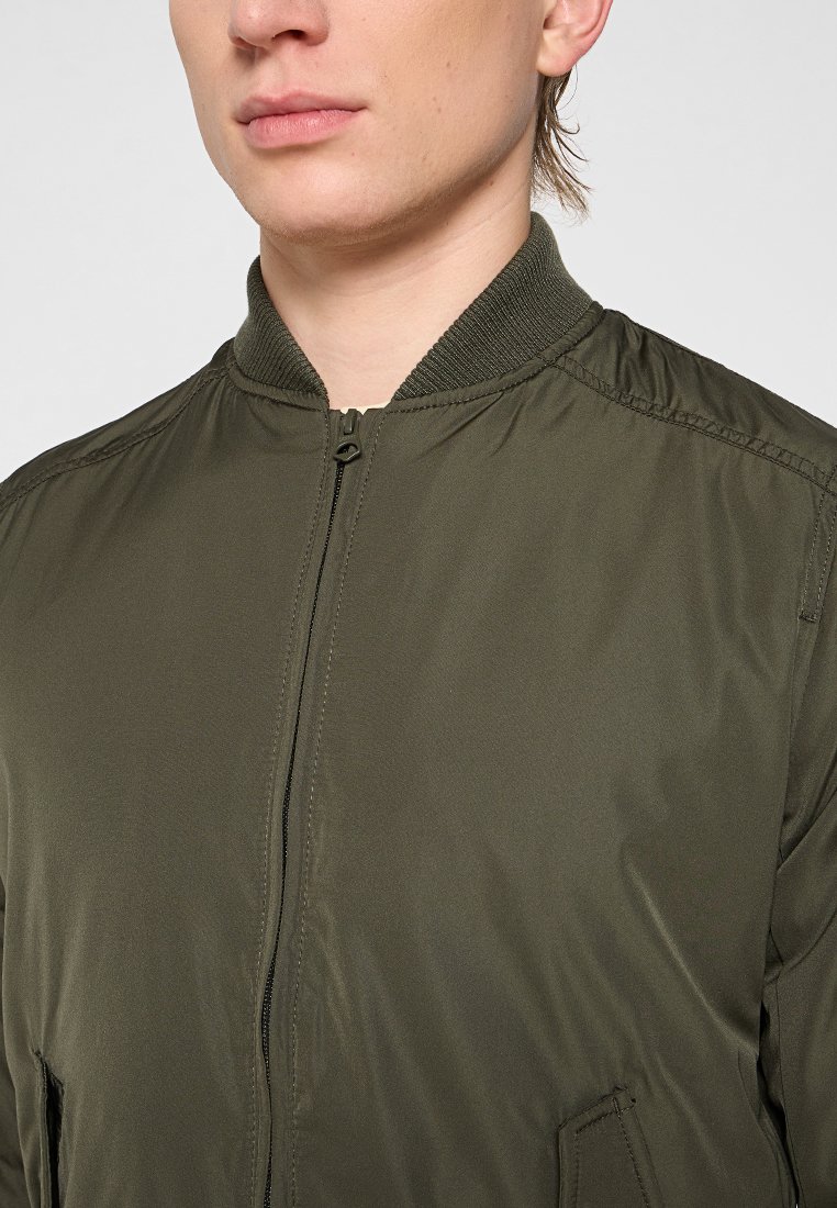 Veste bomber olive avec une fermeture éclair sur le devant, un col côlé et des poches latérales. Texture de tissu lisse avec des détails de couture minimalistes.