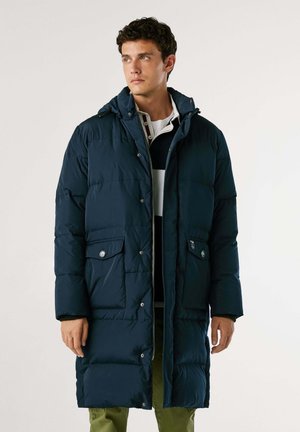 Cappotto imbottito blu navy con cappuccio, bottoni a pressione frontalmente, due tasche anteriori e texture trapuntata. Indossato sopra una maglietta a righe e pantaloni verdi.