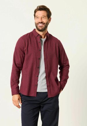 Lächelnder Mann mit kurzem braunem Haar und Bart trägt ein burgunderrotes Hemd über einem grauen T-Shirt und dunklen Hosen, steht mit einer Hand in der Tasche.