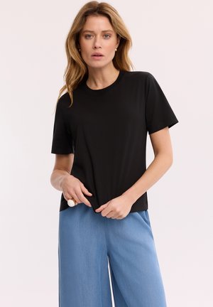 Kobieta o falowanych jasnych włosach, ubrana w prostą czarną koszulkę z krótkim rękawem i luźne jasnoniebieskie spodnie, trzymająca dół koszulki obiema rękami.