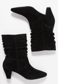 Un par de botas de ante negro a media pantorrilla con caña caída, punta afilada y tacón bajo y grueso, exhibidas sobre un fondo blanco.