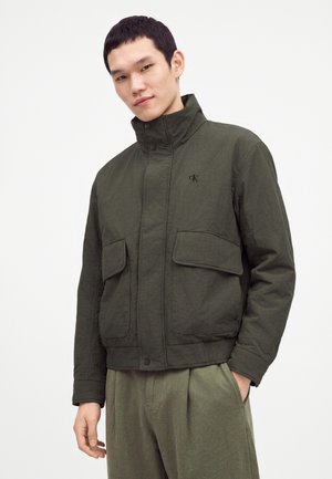 Jeune homme portant une veste vert foncé avec des poches à rabat et un col montant, debout avec une main dans la poche sur un fond uni.