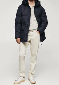 Granatowa kurtka puchowa z kapturem, przednimi kieszeniami i pikowanym wzorem; zestawiona z jasnobeżowymi spodniami i białymi trampkami z beżowymi podeszwami.