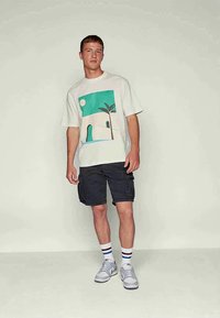 Jovem vestido com uma t-shirt gráfica branca com um design de palmeira, calções de carga pretos, meias listradas e sapatilhas brancas, contra um fundo simples.