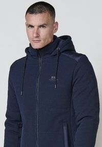 Felpa blu navy con zip in tessuto testurizzato, cappuccio con coulisse e pannelli spalle lisci a contrasto. Presenta un piccolo logo sul petto.