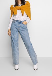 Blusa bianca con pois abbinata a jeans leggermente oversize a vita alta di colore azzurro chiaro. Un maglione lavorato a maglia giallo senape drappeggiato sulle spalle. Stivali bianchi.