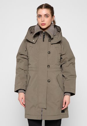 Frau trägt eine taupefarbene Parka mit hohem Kragen, Knöpfen, Reißverschluss und Seitentaschen, steht vor einem schlichten hellgrauen Hintergrund.