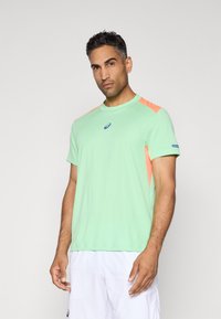 Bărbat îmbrăcat cu un tricou sport de culoare verde deschis, cu accente portocalii, și pantaloni scurți albi, stând în fața unui fundal gri deschis simplu.