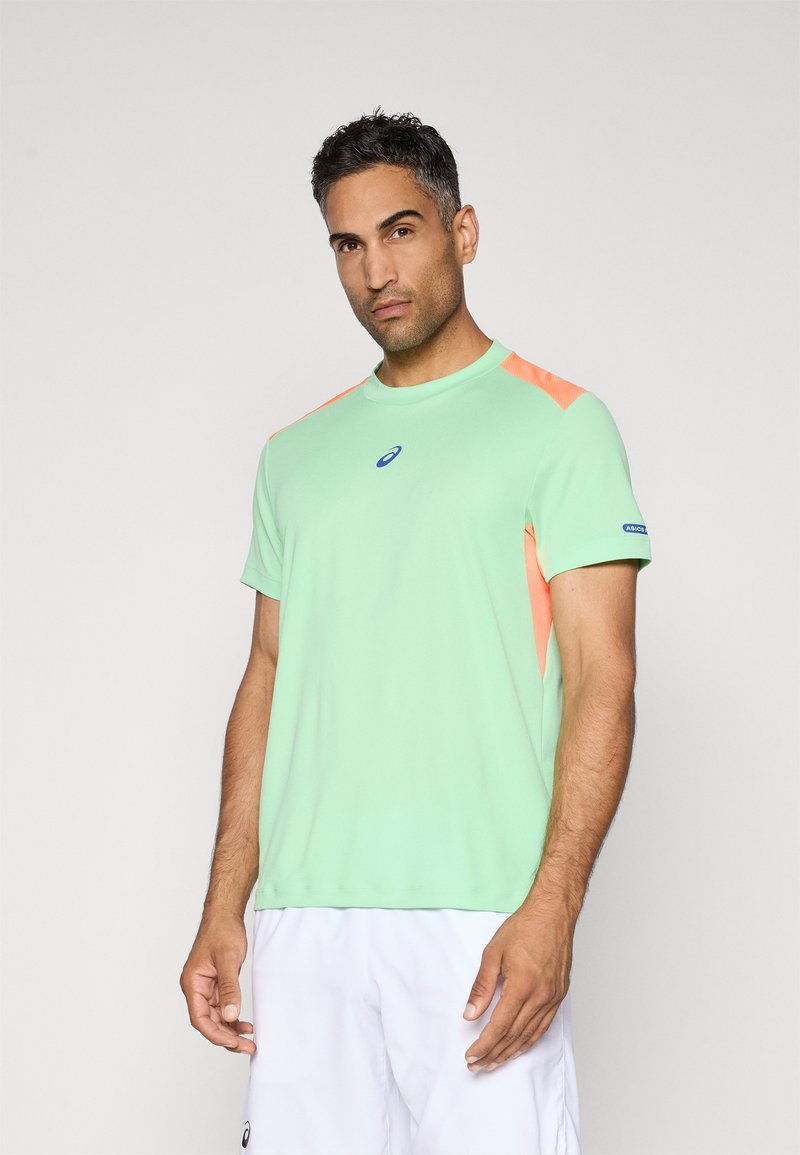 Bărbat îmbrăcat cu un tricou sport de culoare verde deschis, cu accente portocalii, și pantaloni scurți albi, stând în fața unui fundal gri deschis simplu.