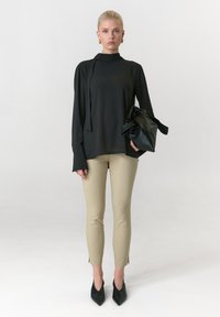 Schwarzes langärmeliges Oberteil mit hohem Hals und seitlicher Schleife, beigefarbene eng anliegende Hose mit Schlitzen an den Knöcheln und spitze schwarze High Heels. Schwarze Handtasche.