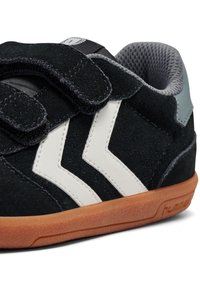 Svart mockaskor med vita zigzag-detaljer, grå mesh-foder och tan gummisula. Har kardborreband för stängning.