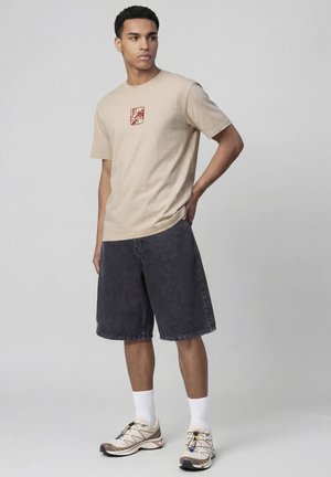 Jeune homme debout portant un t-shirt graphique beige, un short gris foncé mi-cuisses, des chaussettes blanches et des baskets beige sur fond gris.