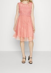 Vestido rosa con cuerpo de encaje y falda transparente. Diseño sin mangas, de longitud a la rodilla, con textura en capas. Combinado con sandalias negras de tacón alto.