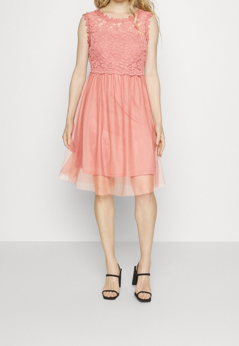 Vestido rosa con cuerpo de encaje y falda transparente. Diseño sin mangas, de longitud a la rodilla, con textura en capas. Combinado con sandalias negras de tacón alto.