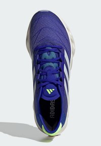 Scarpa sportiva viola con tomaia in rete testurizzata, strisce bianche, dettagli blu e una linguetta sul tallone di colore verde neon. Presenta una linguetta imbottita e lacci.