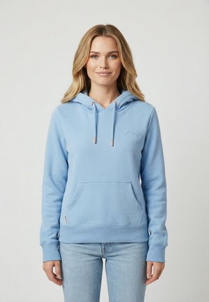 Donna con lunghi capelli biondi che indossa una felpa con cappuccio azzurro chiaro con tasca frontale e jeans azzurri chiari, in piedi davanti a uno sfondo uniforme.