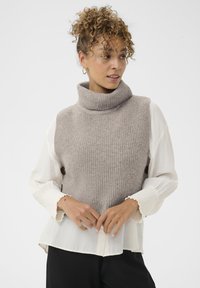 Világosbarna bordázott mintájú kötött garbó mellény, fehér blúzra rétegezve, fodros mandzsettákkal. A modell összekulcsolt kézzel áll elöl.