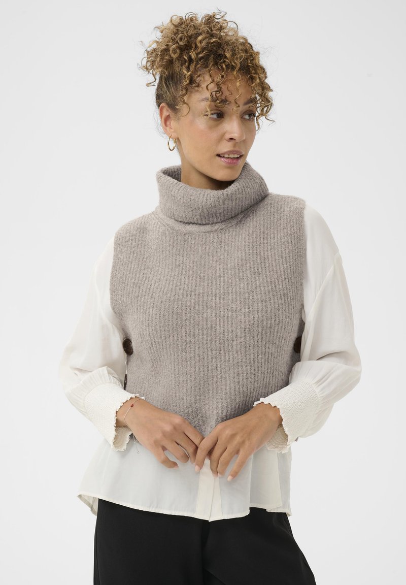 Világosbarna bordázott mintájú kötött garbó mellény, fehér blúzra rétegezve, fodros mandzsettákkal. A modell összekulcsolt kézzel áll elöl.